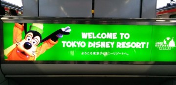 Foto de uma placa de boas vindas com o Pluto, na chegada de metrô na Disney de Tóquio. Ela é verde-limão e tem o texto "Welcome to Tokyo Disney Resort"