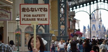 Foto de uma funcionária segurando uma placa em japonês e inglês ("for safety please walk") na Disney de Tóquio, com o castelo ao fundo e visitantes passando