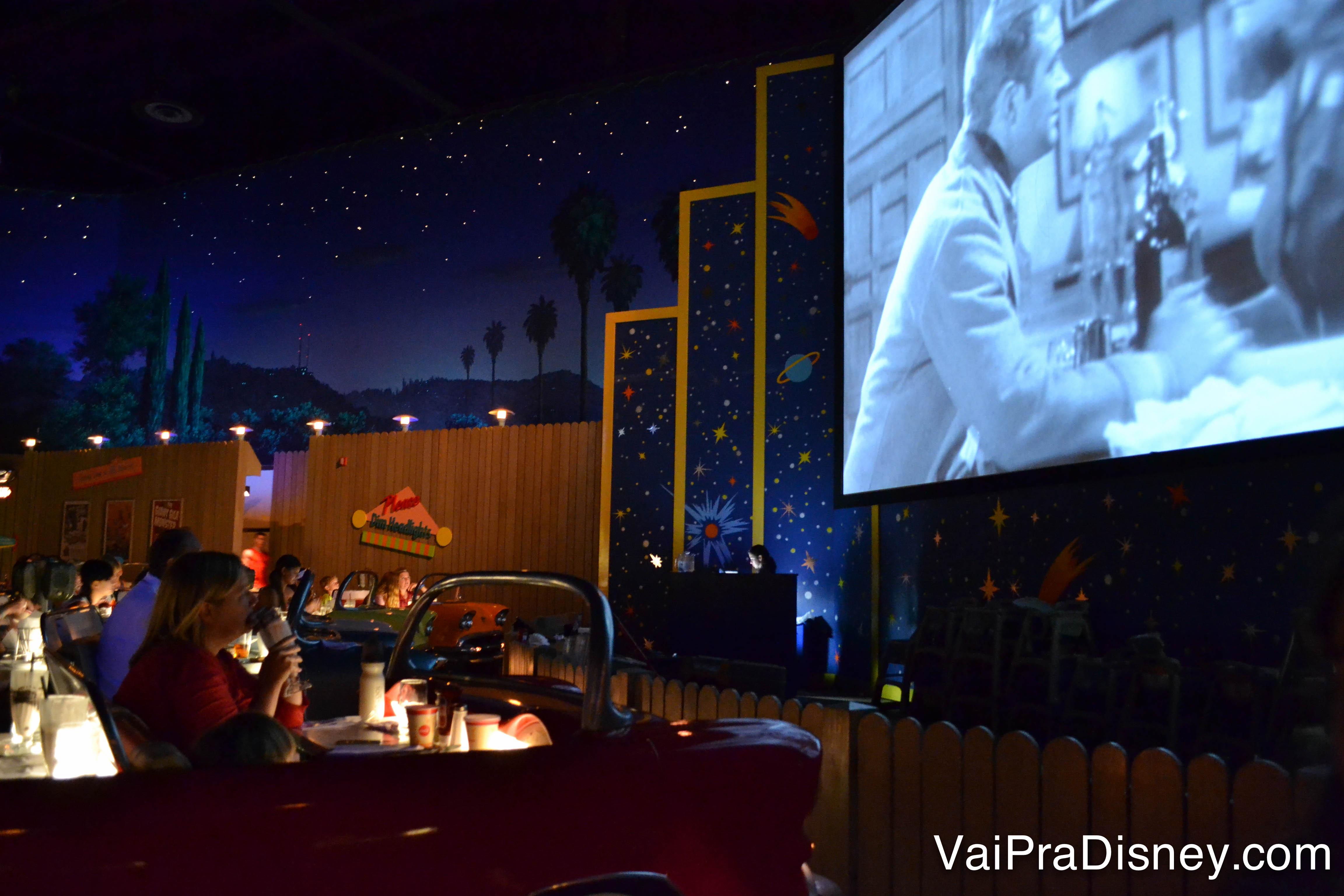 Foto do restaurante, com os carros antigos e a tela passando um filme antigo. O ambiente é escuro para imitar um drive-in à noite 