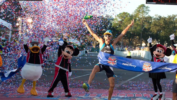 A imagem mostra uma pessoa comemorando por ter terminado a maratona da Disney, ao lado do Mickey, da Minnie e do Donald vestidos com roupas esportivas.