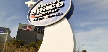 Foto da placa na entrada do Kennedy Space Center, que tem o logo do local e um foguete branco ao lado.