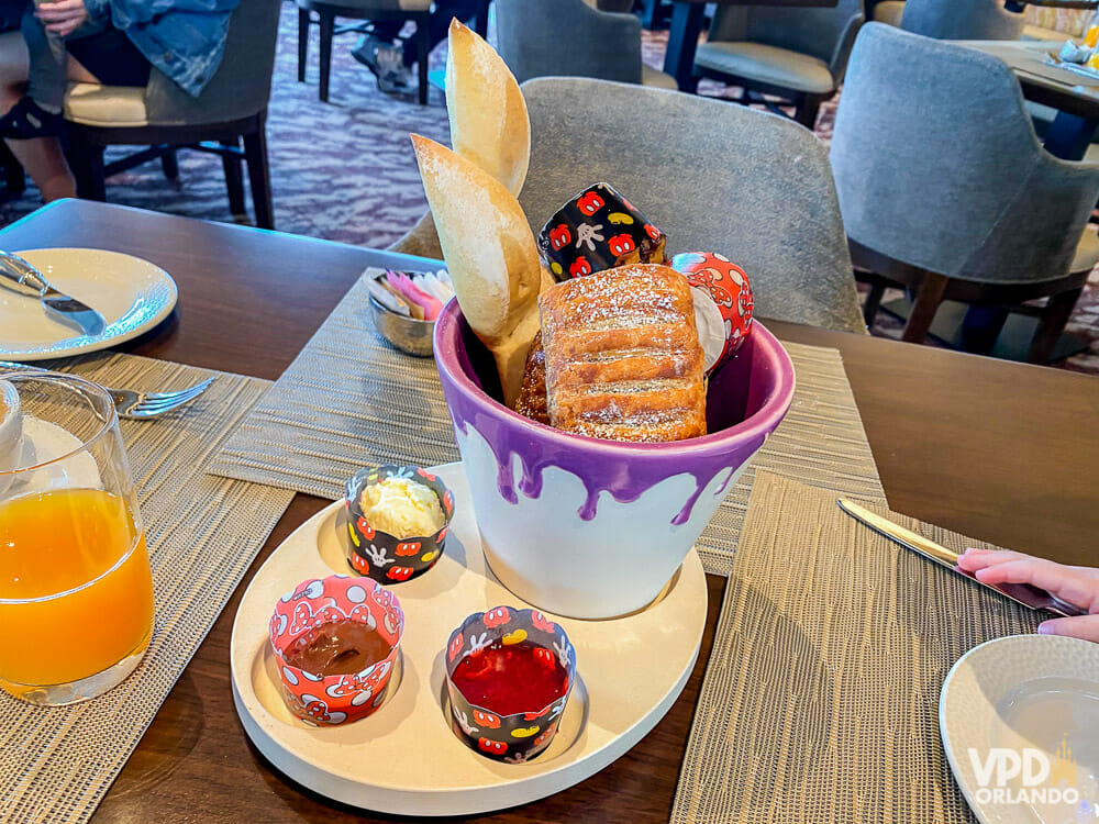 Foto de um prato com uma refeição de um restaurante da Disney, com uma cesta com pães doces e coberturas ao lado