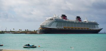 Foto do navio de cruzeiro da Disney Cruise Line perto da praia no Caribe, com o mar azul-turquesa, a areia clara e o céu claro atrás.