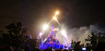 Foto do castelo da Bela Adormecida da Disneyland de Paris durante o show de fogos. O castelo está iluminado em azul e rosa, e o céu está cheio de fogos de artifício explodindo.