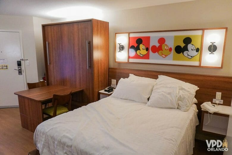 Imagem do quarto reformado do hotel Pop Century. Há uma cama de casal com lençóis brancos e um quadro com três Mickeys em cores diferentes.