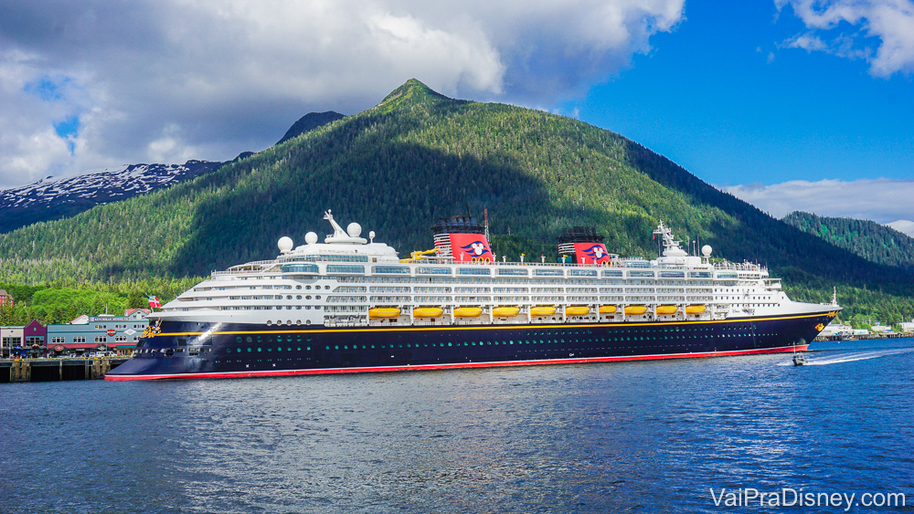 Foto do navio de cruzeiro da Disney, o Disney Magic, em alto mar, com montanhas e o céu azul ao fundo. Ele é pintado de azul marinho com detalhes em vermelho e amarelo, e as chaminés imitam a roupa do Mickey.