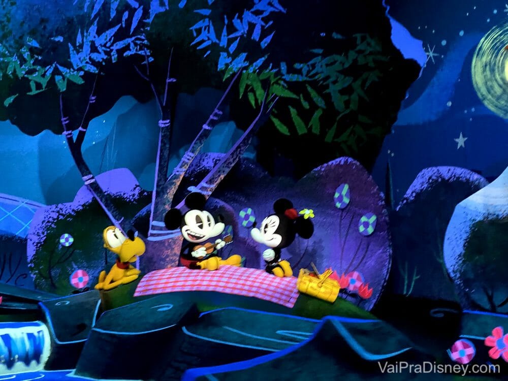 Imagem de divulgação da atração Mickey & Minnie's Runaway Railway, que mostra o Mickey, a Minnie e o Pluto fazendo um piquenique à noite sob árvores.