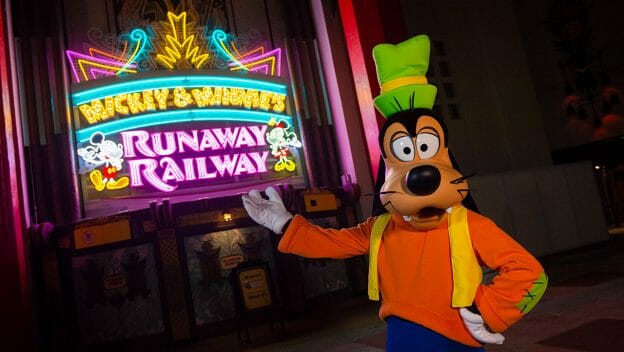 Imagem do Pateta apontando para o Teatro Chinês, onde há um letreiro luminoso com o nome da nova atração do Mickey e da Minnie, Mickey & Minnie's Runaway Railway