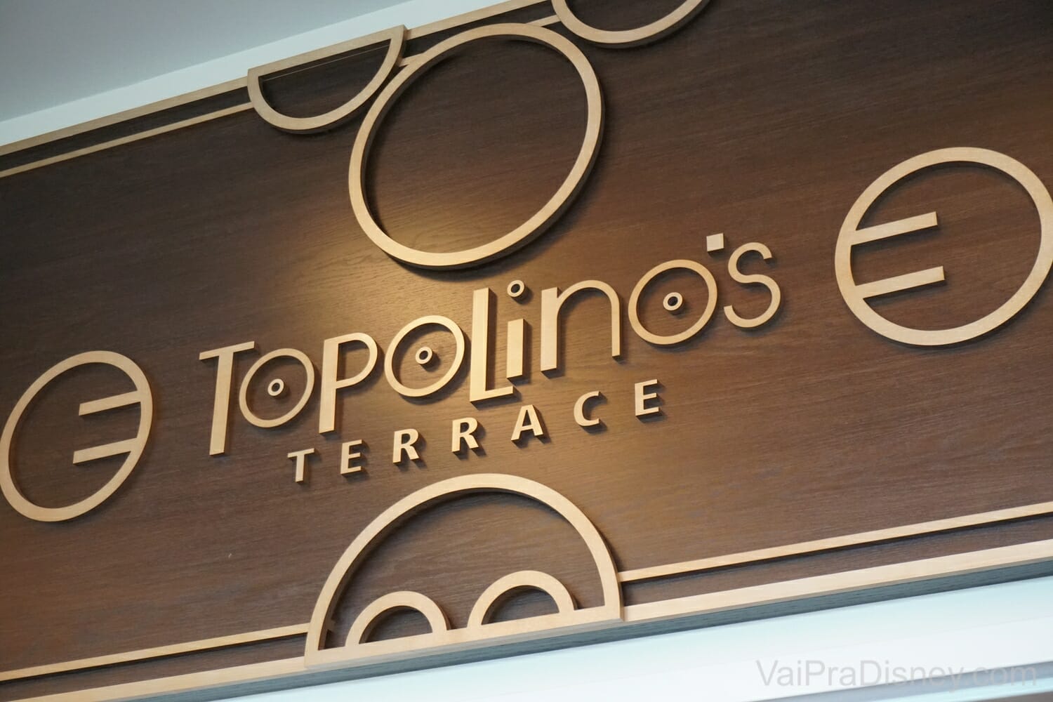 Foto da placa na entrada do restaurante Topolino's Terrace, do Disney's Riviera Resort. A placa é marrom e tem as orelhas, mãos e os botões da calça do Mickey.