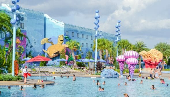 A imagem mostra visitantes na piscina do hotel da Disney, o Art of Animation. A decoração é colorida, o prédio ao fundo é azul e a tartaruga do filme Procurando Nemo é parte da decoração.
