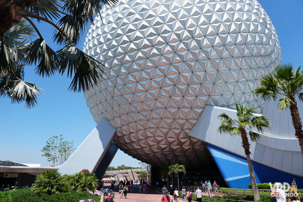 Foto da "bola" que é símbolo do Epcot, onde fica a atração Spaceship Earth, com o céu azul ao fundo e alguns visitantes passando ao redor.