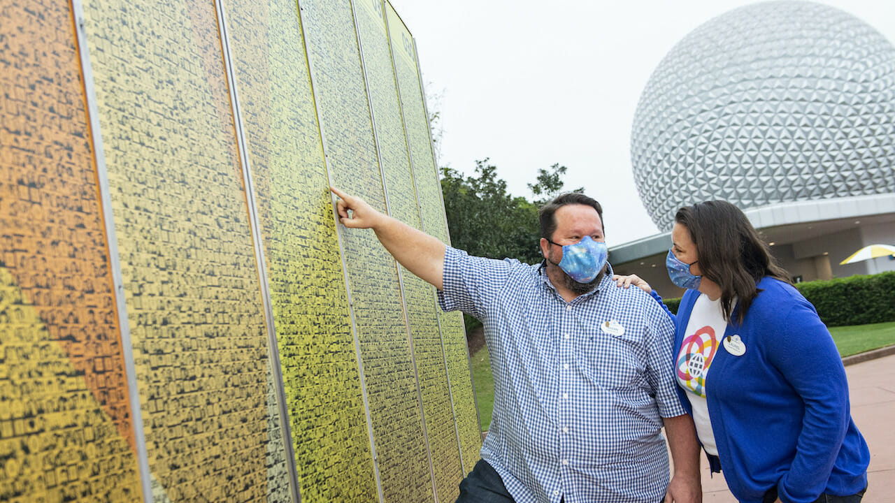 Imagem de dois visitantes em frente aos novos monolitos do Leave a Legacy, com a Spaceship Earth ao fundo. Ambos usam roupas azuis e usam máscaras também azuis.